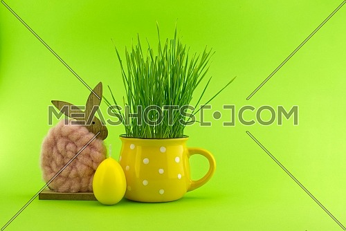 Easter Decoration with wheat seedlings growing from yellow cup, egg shaped candle and Easter Rabbit figure over a green background with free copy space for text