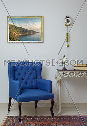 Vintage Furniture - Interior composition of retro blue armchair, vintage wooden beige table, table lamp, books, and pendulum clock over off white wall, tiled beige floor and orange ornate carpet