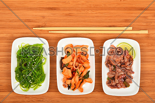 Three portions of seafood marinated salad with octopus cuttlefish, squid and seaweeds in small white plates with chopsticks on bamboo wood, top view