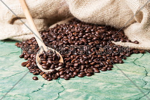 Coffee beans in coffee burlap bag on green surface and wooden spoon with coffee beans on top.
