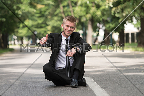 Happy Businessman Sitting on Asphalt Outdoors In Park And Showing Thumbs-Up Sign