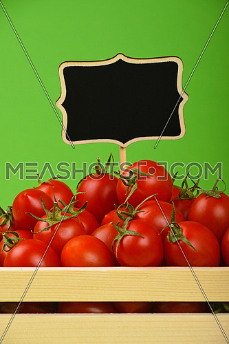 Fresh red ripe cherry tomatoes in small wooden box with black chalkboard price sign tag over green background