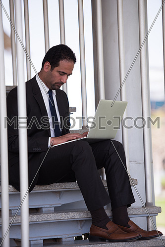 Young handsome middle eastern business executive sitting on steps in a corporate offices building using laptop