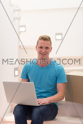 Happy young man sitting in stairway at home, using laptop computer with boxes around him