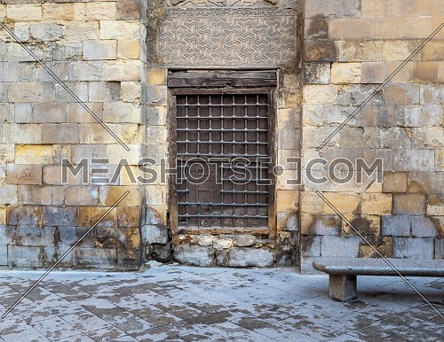 Wooden window with decorated iron grid over stone bricks wall and marble garden bench, Moez Street, Cairo, Egypt