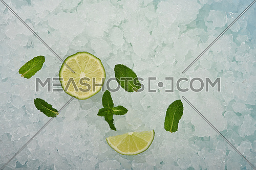Close up fresh juicy green lime cut half and wedge slice and mint leaves over background of crushed ice, elevated high angle view, directly above