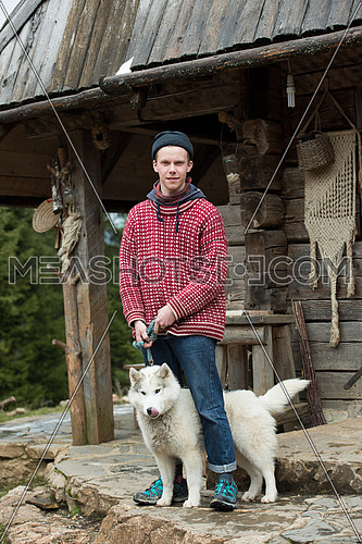 handsome young hipster portrait, young  man standing together with white husky dog in front of old vintage retro wooden house