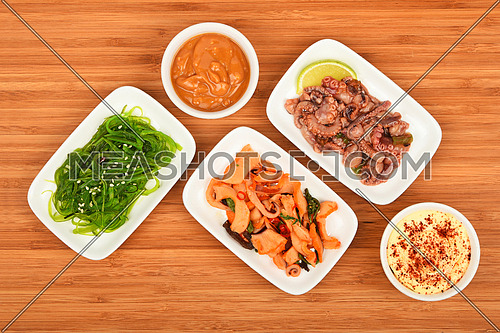 Three portions of seafood marinated salad with octopus cuttlefish, squid and seaweeds in small white plates with sauce on wooden table, top view