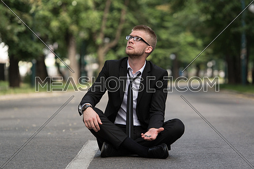 Young Businessman Sitting on Asphalt Begs For Money Outdoors In Park